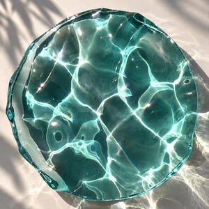 May include: A circular teal glass dish featuring a textured surface that mimics the appearance of water ripples. The glass is translucent, creating light reflections and shadows. The text OCEAN WATER RIPPLE ART is printed above the dish.