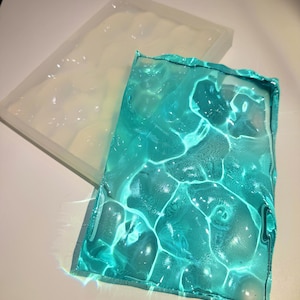 May include: A clear silicone mould with a wavy pattern is next to a turquoise resin piece with a water effect. The resin piece has a rippled surface, creating a water-like appearance. The items are on a white surface.