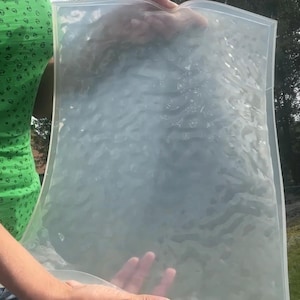 Puede incluir: Un molde rectangular de silicona translúcida con una superficie texturizada y ondulada que imita la apariencia de agua en movimiento. El material flexible y transparente tiene un borde elevado liso y una textura interior suave e irregular.