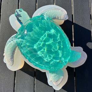 Puede incluir: Una escultura de tortuga marina de vidrio azul verdoso con una concha de resina blanca. La tortuga está sentada sobre una superficie de madera negra.