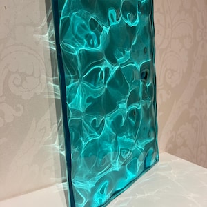 May include: A rectangular, teal-colored glass panel with a textured surface, creating a rippled water effect. The glass is illuminated, casting light and shadow patterns. The panel is displayed on a white surface.