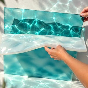 Può includere: Un pannello rettangolare in vetro color verde acqua con una texture superficiale ondulata simile all'acqua che crea motivi di rifrazione della luce. Uno stampo in silicone bianco traslucido e flessibile con bordo ondulato è posizionato.