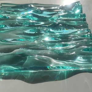 Pode incluir: Uma placa de vidro retangular de cor verde-azulado translúcida com uma superfície texturizada que imita a aparência de água ondulante. O vidro tem cristas e vales elevados que captam a luz, criando reflexos brilhantes.