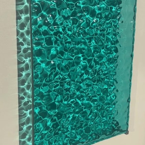 May include: A rectangular wall-mounted panel featuring a translucent teal glass surface with a rippled, water-like texture. The glass is encased in a thin, metallic silver frame that holds the textured sheet, creating light refractions.