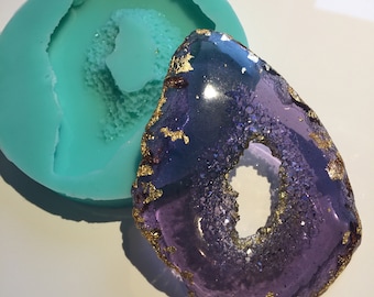 May include: A purple and gold agate slice with a hole in the center, sitting on a teal silicone mold.
