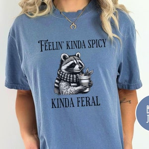 以下が含まれることがあります： 青いジーンズのTシャツに、スカーフを巻いてコーヒーカップを持ったアライグマが描かれています。Tシャツには「Feelin' Kinda Spicy Kinda Feral」と書いてあります。