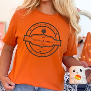 May include: Orange t-shirt with a circular graphic that reads "Three Witches Brewing Company" in a dark gray color. The shirt is short-sleeved and has a crew neck. A white mug with a ghost design and candy corn is held in the hand.