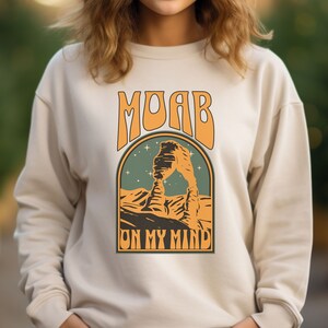 May include: A cream-colored sweatshirt with a retro-style graphic of a red rock arch with the text "MOAB ON MY MIND" in orange.