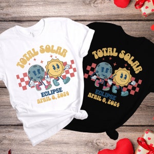 May include: Two white and black t-shirts with a retro design featuring a cartoon sun and moon with the text "Total Solar Eclipse April 8, 2024".