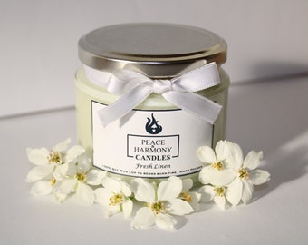 May include: A white candle jar with a silver lid and a white ribbon tied in a bow. The label reads "Peace & Harmony Candles" and "Fresh Linen".