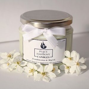 May include: A glass jar candle with a silver lid and a white ribbon tied around the neck. The label reads "Peace Harmony Candles, Frankincense & Myrrh." White flowers surround the candle. The candle is hand-poured and made with soy wax.
