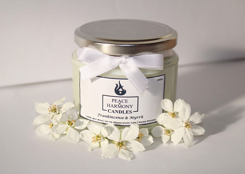 May include: A glass jar candle with a silver lid and a white ribbon tied around the neck. The label reads "Peace Harmony Candles, Frankincense & Myrrh." White flowers surround the candle. The candle is hand-poured and made with soy wax.
