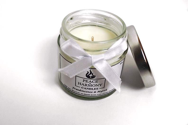 May include: A small glass jar candle with a white wax interior and a silver lid. The jar is adorned with a white ribbon and a label that reads "Peace Harmony Candles" with the scent "Frankincense & Myrrh".