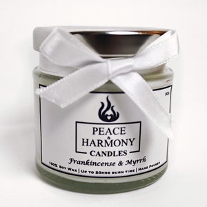 May include: A small glass jar candle with a silver lid and a white ribbon tied in a bow. The label reads "Peace Harmony Candles" with "Frankincense & Myrrh" below. The candle is filled with a pale green wax.