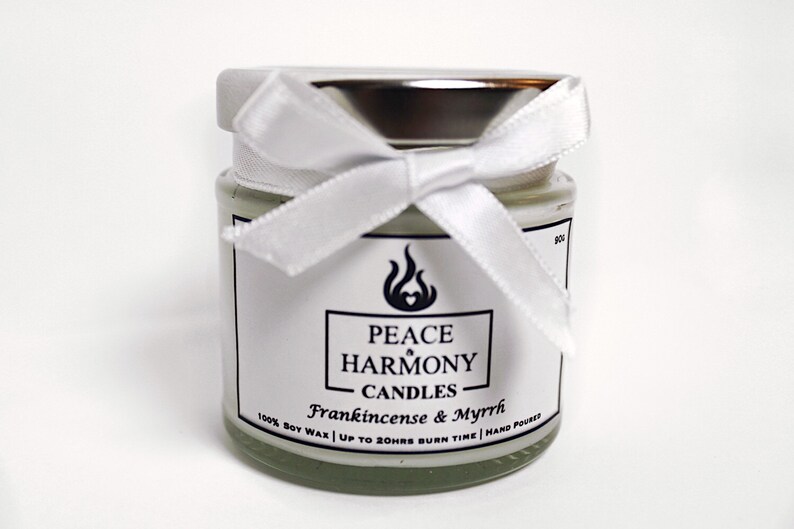 May include: A small glass jar candle with a silver lid and a white ribbon tied in a bow. The label reads "Peace Harmony Candles" with "Frankincense & Myrrh" below. The candle is filled with a pale green wax.