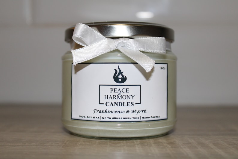 May include: A glass jar candle with a silver lid and a white ribbon tied around the top. The label reads "Peace & Harmony Candles" with "Frankincense & Myrrh" below. The candle is filled with a pale green wax.