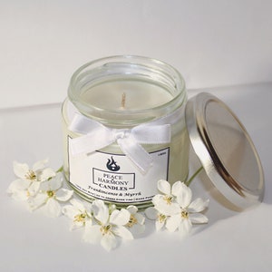 May include: A clear glass jar candle with a silver lid and a white ribbon tied around the jar. The candle has a white label with the text "Peace Harmony Candles" and "Frankincense & Myrrh". White flowers are arranged around the candle.