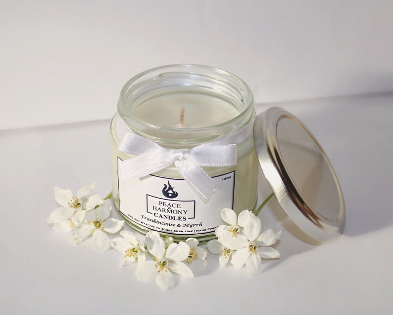 May include: A clear glass jar candle with a silver lid and a white ribbon tied around the jar. The candle has a white label with the text "Peace Harmony Candles" and "Frankincense & Myrrh". White flowers are arranged around the candle.