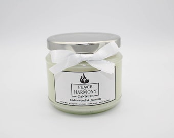May include: A white candle in a glass jar with a silver lid. The jar is tied with a white ribbon and has a label that reads "Peace Harmony Candles - Cedarwood & Jasmine".