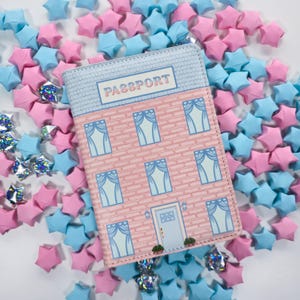 May include: A passport cover featuring a pink brick design, a blue roof, and the word "PASSPORT". The cover is surrounded by pink and blue origami stars. The design resembles a house.
