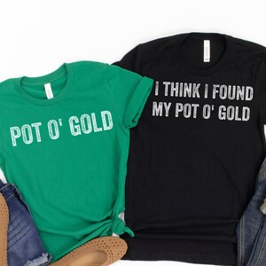 May include: Two t-shirts with a St. Patrick's Day theme. One shirt is green and says "POT O' GOLD" in white text. The other shirt is black and says "I THINK I FOUND MY POT O' GOLD" in white text.