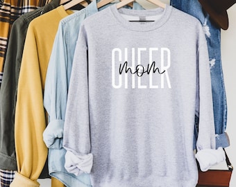 May include: A grey sweatshirt with the text "CHEER mom" printed in black and white on the front. The sweatshirt is hanging on a rack with other clothing items.