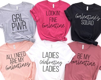 May include: Six different t-shirts with various Galentine's Day themed slogans. The shirts are in different colors, including pink, white, black, gray, and red. The slogans include "GRL PWR", "Lookin' Fine Galentine", "Galentine's Squad", "All I Need Are My Galentines", "Ladies Celebrating Ladies", and "Be My Galentine".