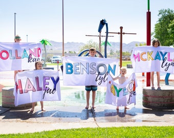 May include: Five children hold up colorful towels with their names written on them in a playful font. The towels are white with multi-colored letters. The children are standing in front of a water park with a blue sky in the background.