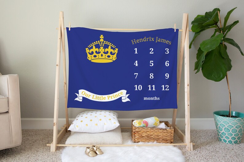 May include: A blue baby milestone blanket with a gold crown and the text "Our Little Prince". The blanket also has a white grid with numbers 1 through 12 for each month of the year and the text "Hendrix James" and "months".