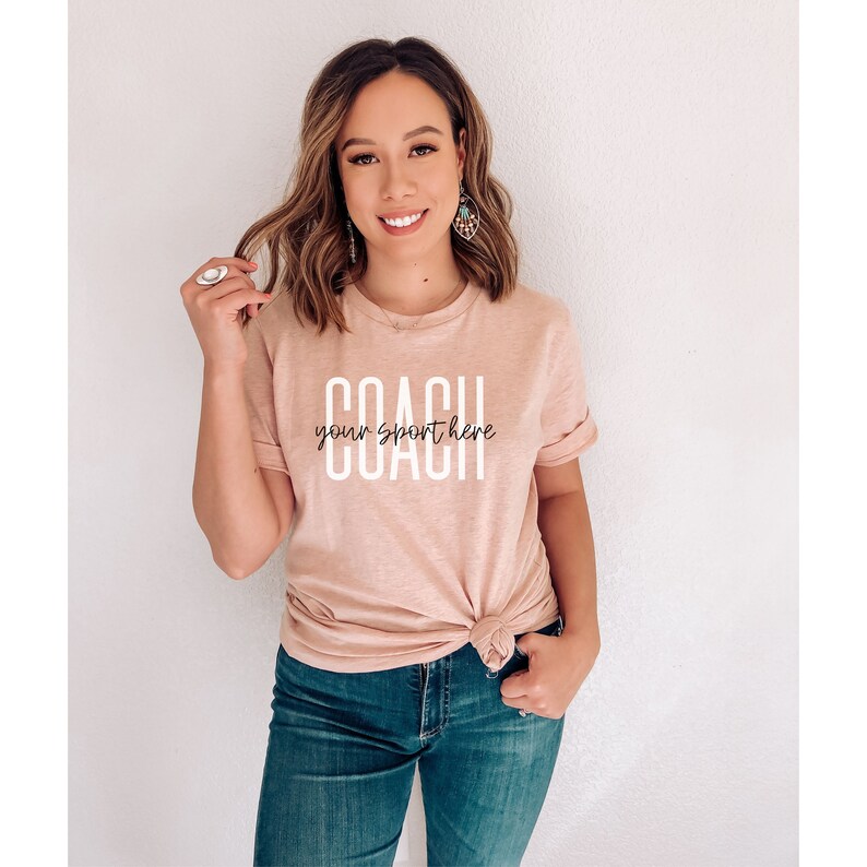 Op de afbeelding: Een lichtroze T-shirt met de tekst "COACH your sport here" in witte letters.