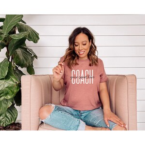 Op de afbeelding: Een vrouw draagt een mauve t-shirt met de tekst "COACH your sport here" in witte letters. Ze zit op een roze bank.