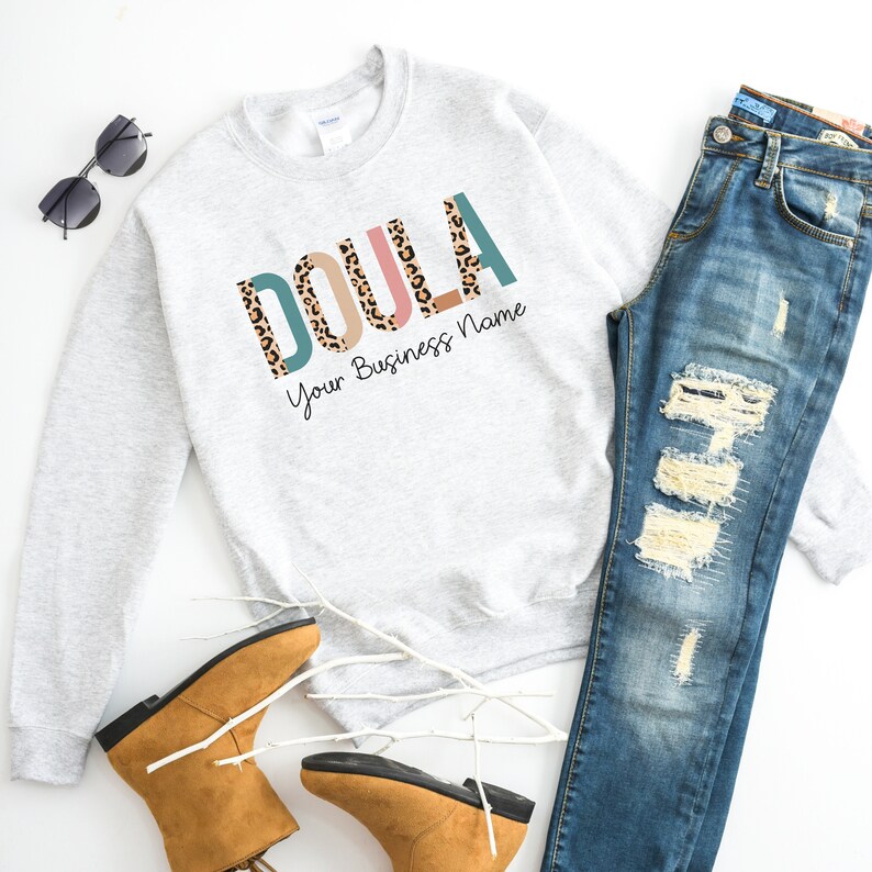 May include: A gray sweatshirt with the word "DOULA" printed in colorful letters with a leopard print pattern. The text "Your Business Name" is printed below the word "DOULA".