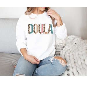 May include: White sweatshirt with the word "DOULA" printed in large letters. The letters are outlined in teal, pink, and brown, and filled with a leopard print pattern.