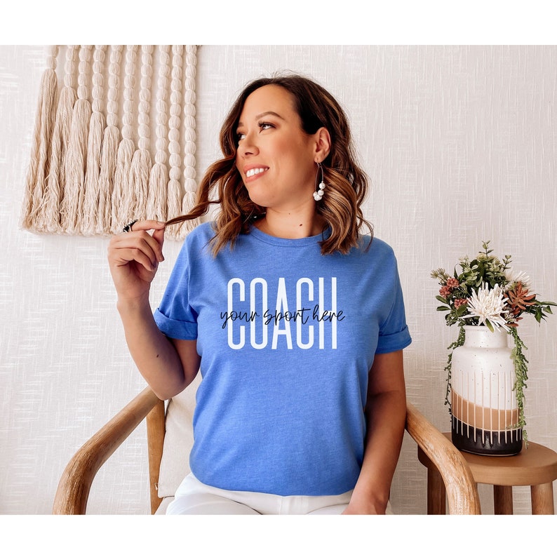 Op de afbeelding: Een blauw t-shirt met witte tekst die "COACH your sport here" luidt.