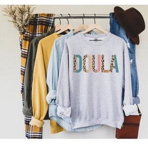 May include: A gray sweatshirt with the word "DOULA" printed in colorful letters with a leopard print pattern. The letters are outlined in teal, pink, and brown.