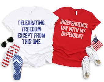 May include: Two white t-shirts with red and blue text. The white shirt on the left says "Celebrating Freedom Except From This One". The red shirt on the right says "Independence Day With My Dependent".