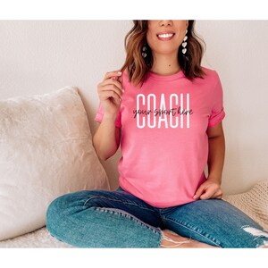 Op de afbeelding: Een roze T-shirt met de tekst "COACH your sport here" in witte letters.