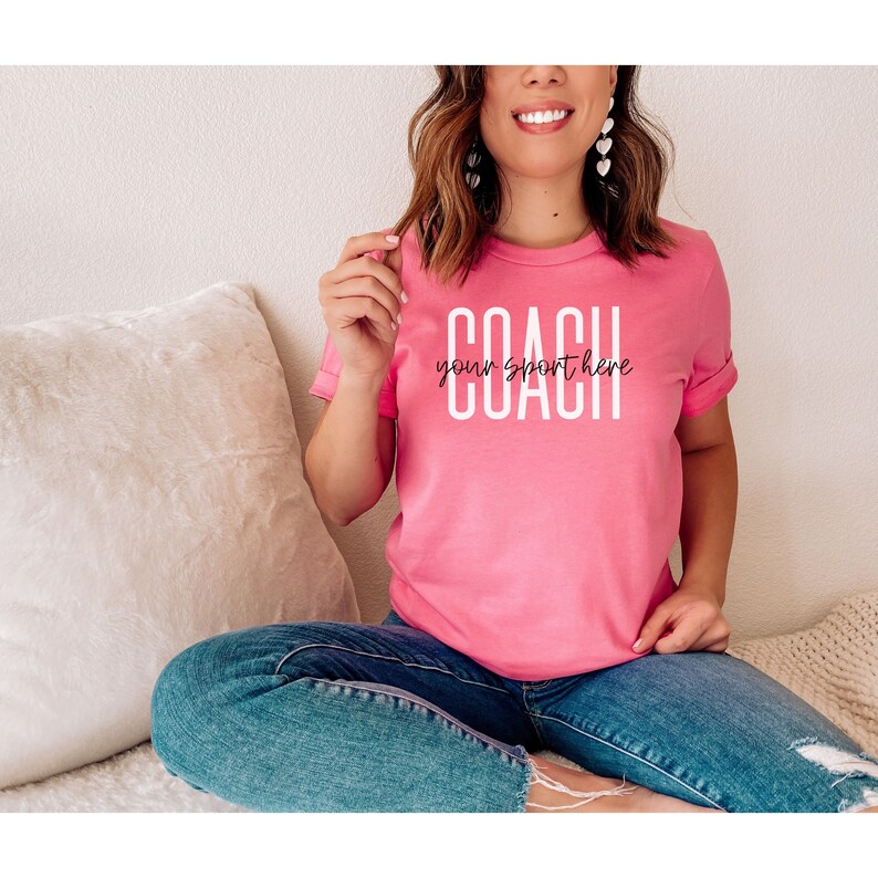 Op de afbeelding: Een roze T-shirt met de tekst "COACH your sport here" in witte letters.