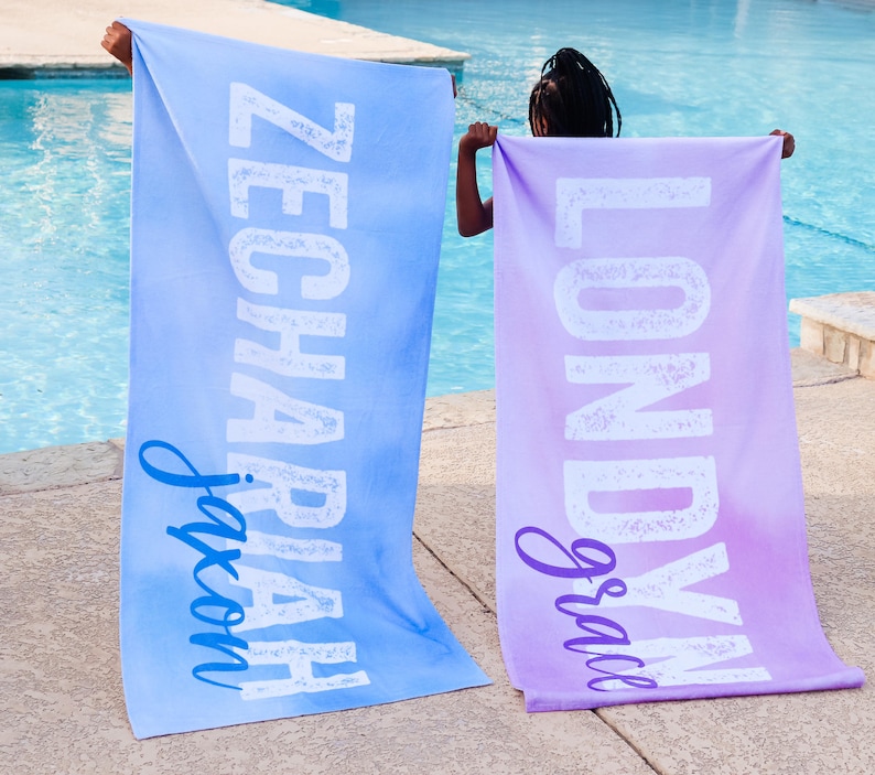 Pode incluir: Duas toalhas de praia personalizadas com os nomes "Jaxon" e "Gracie" impressos em branco sobre um fundo azul e roxo degrad&ecirc;.