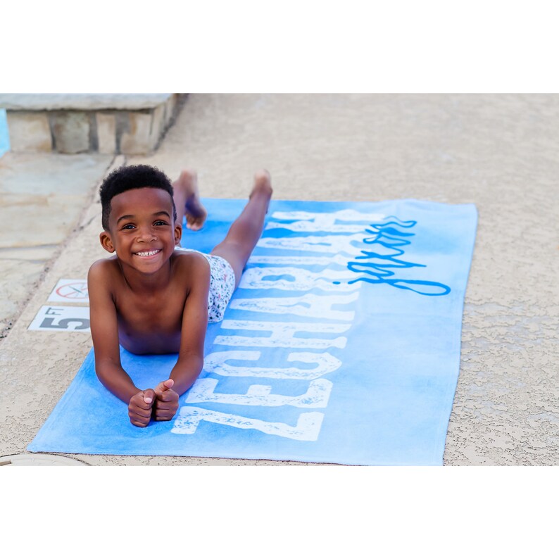 Pode incluir: Um menino pequeno est&aacute; deitado em uma toalha de praia azul com as palavras "Beach Days" impressas em branco. A toalha tem um degrad&ecirc; de cores azuis.