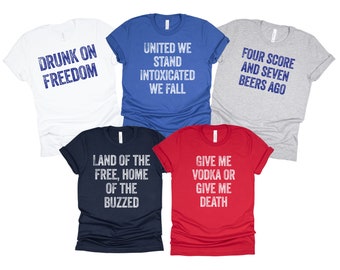 May include: Five t-shirts with humorous, patriotic, and alcohol-themed slogans printed in white on different colored shirts. The slogans are "Drunk on Freedom", "United We Stand Intoxicated We Fall", "Four Score and Seven Beers Ago", "Land of the Free, Home of the Buzzed", and "Give Me Vodka or Give Me Death".