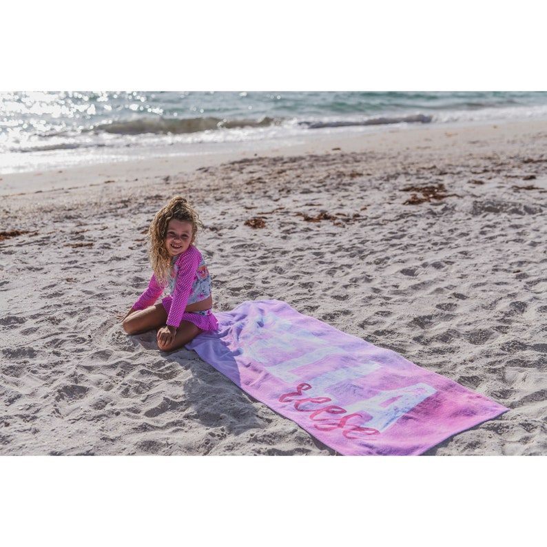 Pode incluir: Uma menina pequena est&aacute; sentada em uma toalha de praia rosa com a palavra "Slesse" escrita nela. A toalha est&aacute; em uma praia de areia com o oceano ao fundo.