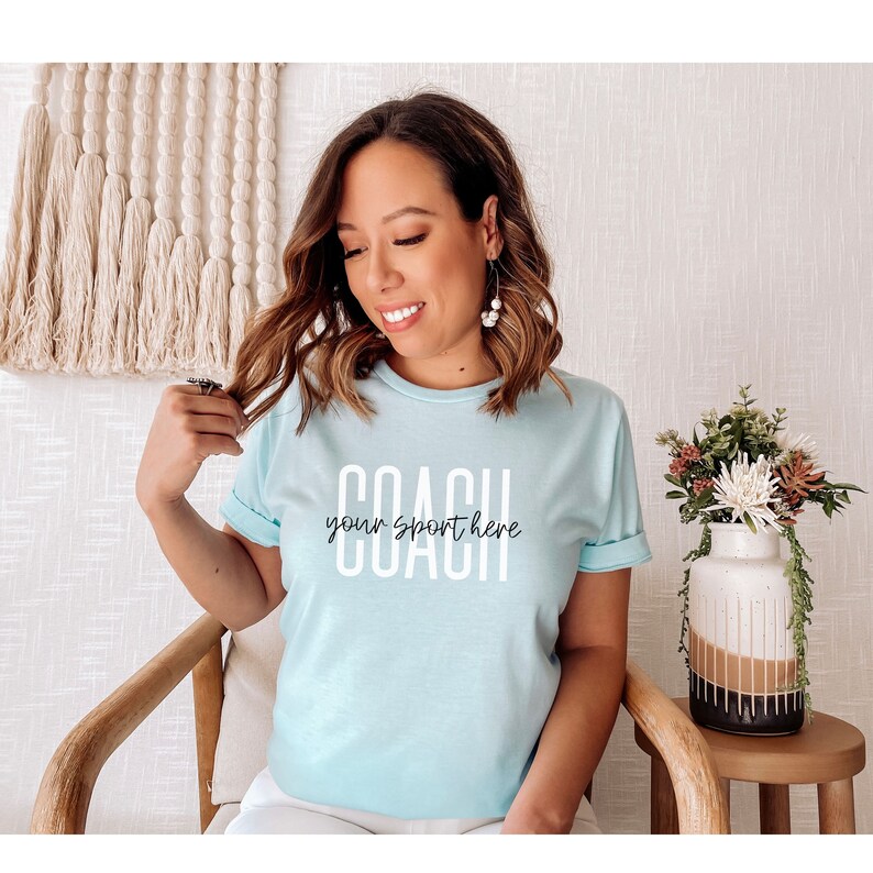 Op de afbeelding: Een lichtblauw t-shirt met de tekst "COACH your sport here" in witte letters. Het shirt wordt gedragen door een persoon met lang bruin haar.