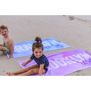 Pode incluir: Duas crian&ccedil;as numa praia, uma sentada numa toalha de praia azul e roxa com a palavra "Brayden" impressa nela, e a outra sentada numa toalha de praia roxa com a palavra "Brooklyn" impressa nela.