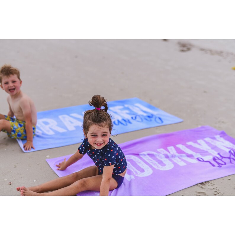 Pode incluir: Duas crian&ccedil;as numa praia, uma sentada numa toalha de praia azul e roxa com a palavra "Brayden" impressa nela, e a outra sentada numa toalha de praia roxa com a palavra "Brooklyn" impressa nela.