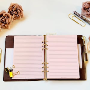 May include: Open brown leather planner with pink lined paper, gold accents, and a diamond-topped pen. Decorative roses, paper clips, and a perfume bottle with the word "ROSE" are arranged around the planner.