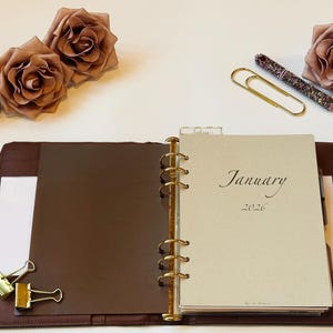 Puede incluir: Un planificador de cuero marrón abierto con la inscripción "Enero 2026". La imagen incluye rosas decorativas, clips, un bolígrafo y un frasco de perfume. El planificador está rodeado de artículos de papelería.