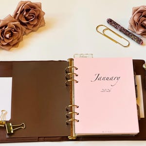May include: Open brown leather planner with gold rings, a pink January 2026 page, and a pen. Gold paper clips, a perfume bottle, and fabric roses are arranged around the planner on a white surface.
