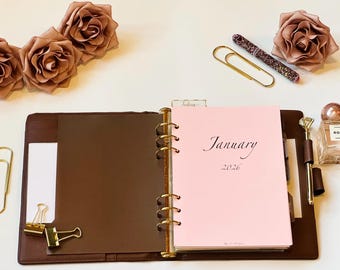 May include: Open brown leather planner with gold rings, a pink January 2026 page, and a pen. Gold paper clips, a perfume bottle, and fabric roses are arranged around the planner on a white surface.