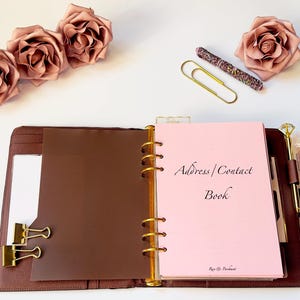 May include: A brown leather binder with gold detailing, open to a pink page with the text "Address | Contact Book." The image includes decorative roses, a pen, paper clips, and a perfume bottle.